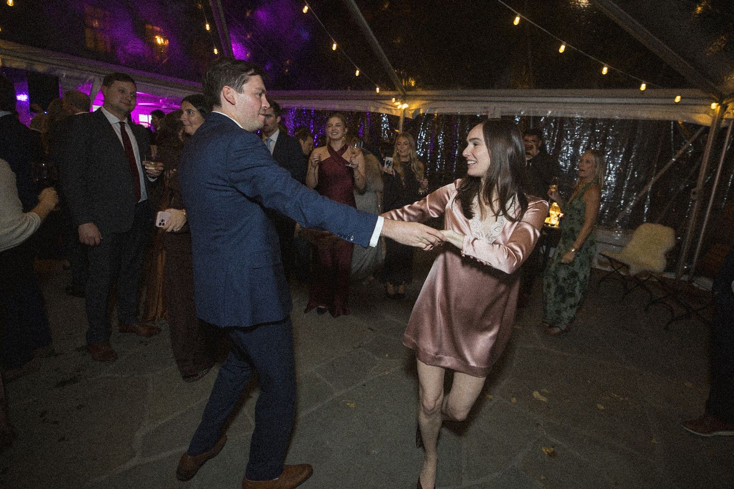 Wedding guests dance and hold hands under a tent with string lights, a woman in a blush satin dress smiling beside a man in a navy suit.