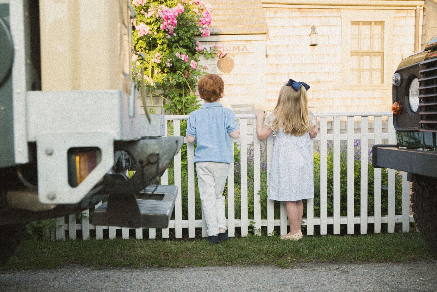 Two children stand by a white picket fence between vintage trucks, girl wearing blue bow, boy in light blue shirt.