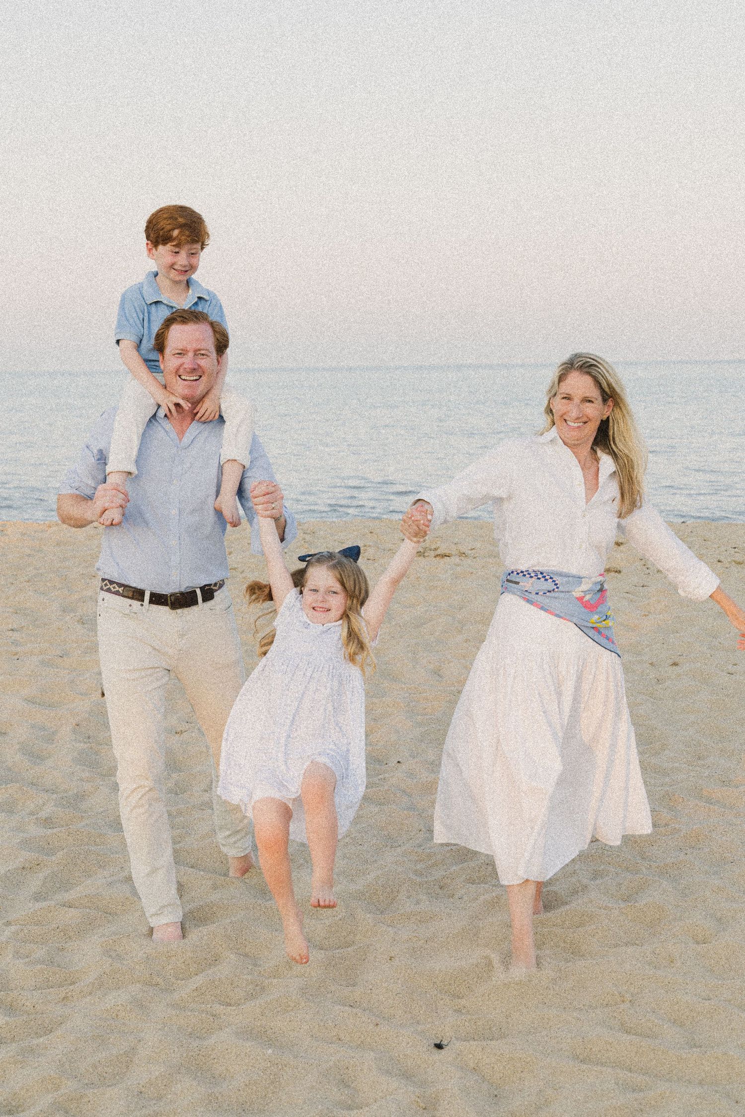 A joyful family of four plays on a sandy beach at sunset, with a boy on dad’s shoulders and a girl held between parents.
