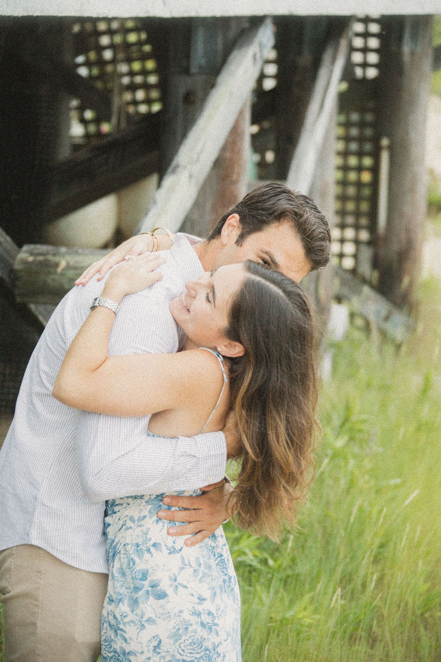 A couple shares a romantic embrace outdoors, with the man kissing the woman near a rustic wooden structure.