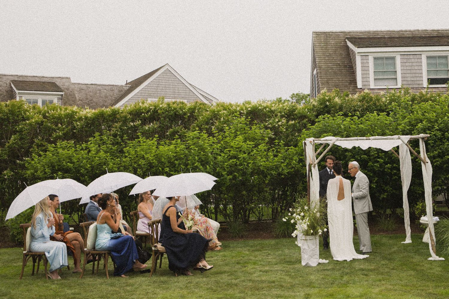 Outdoor wedding ceremony on a rainy day with guests holding white umbrellas seated on lawn chairs near a floral arch.