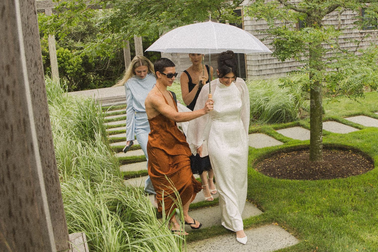 Women walking in a lush garden under a white umbrella on a rainy day, wearing elegant outfits.