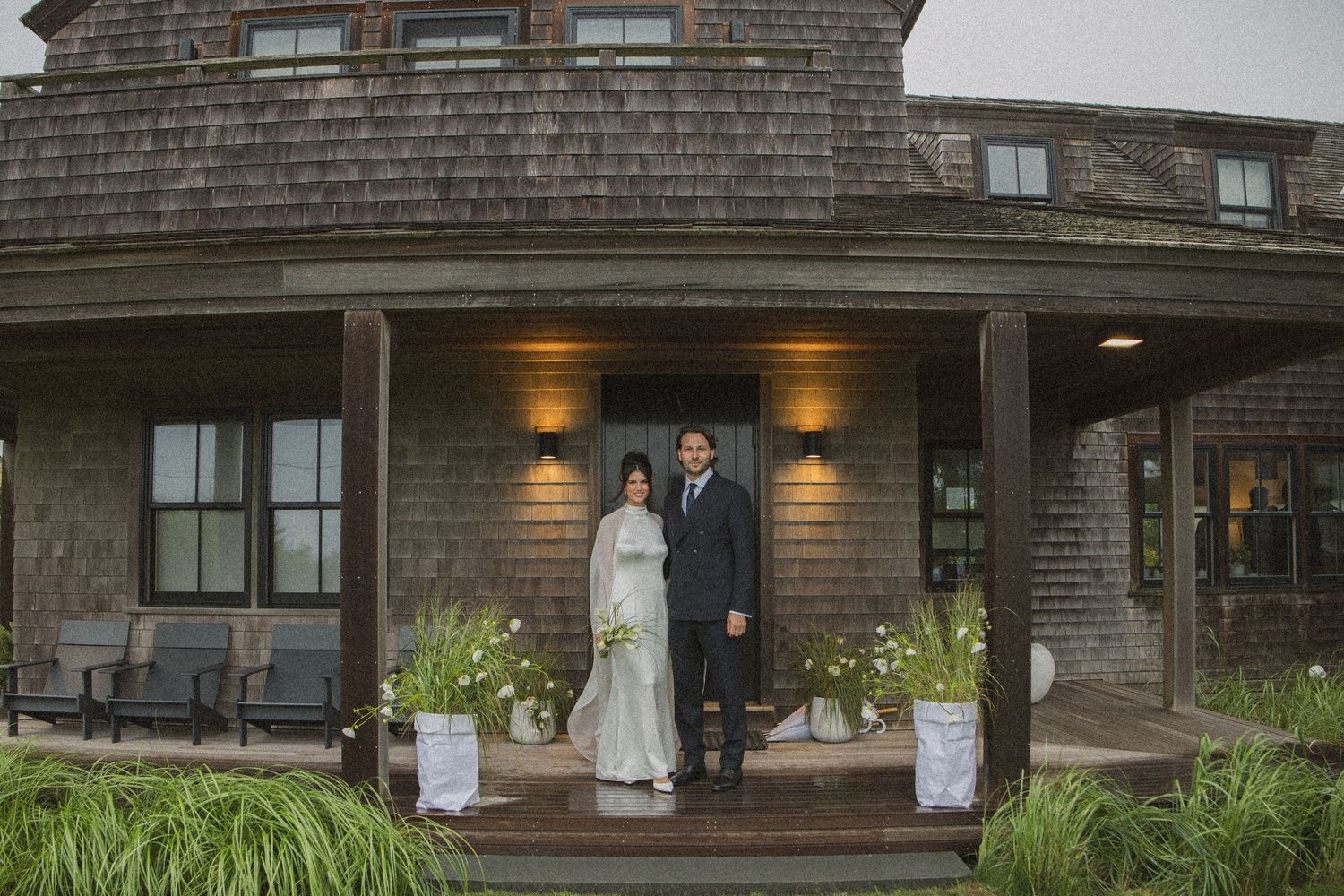 Couple in formal attire standing on porch of rustic shingle-style building surrounded by lush greenery.