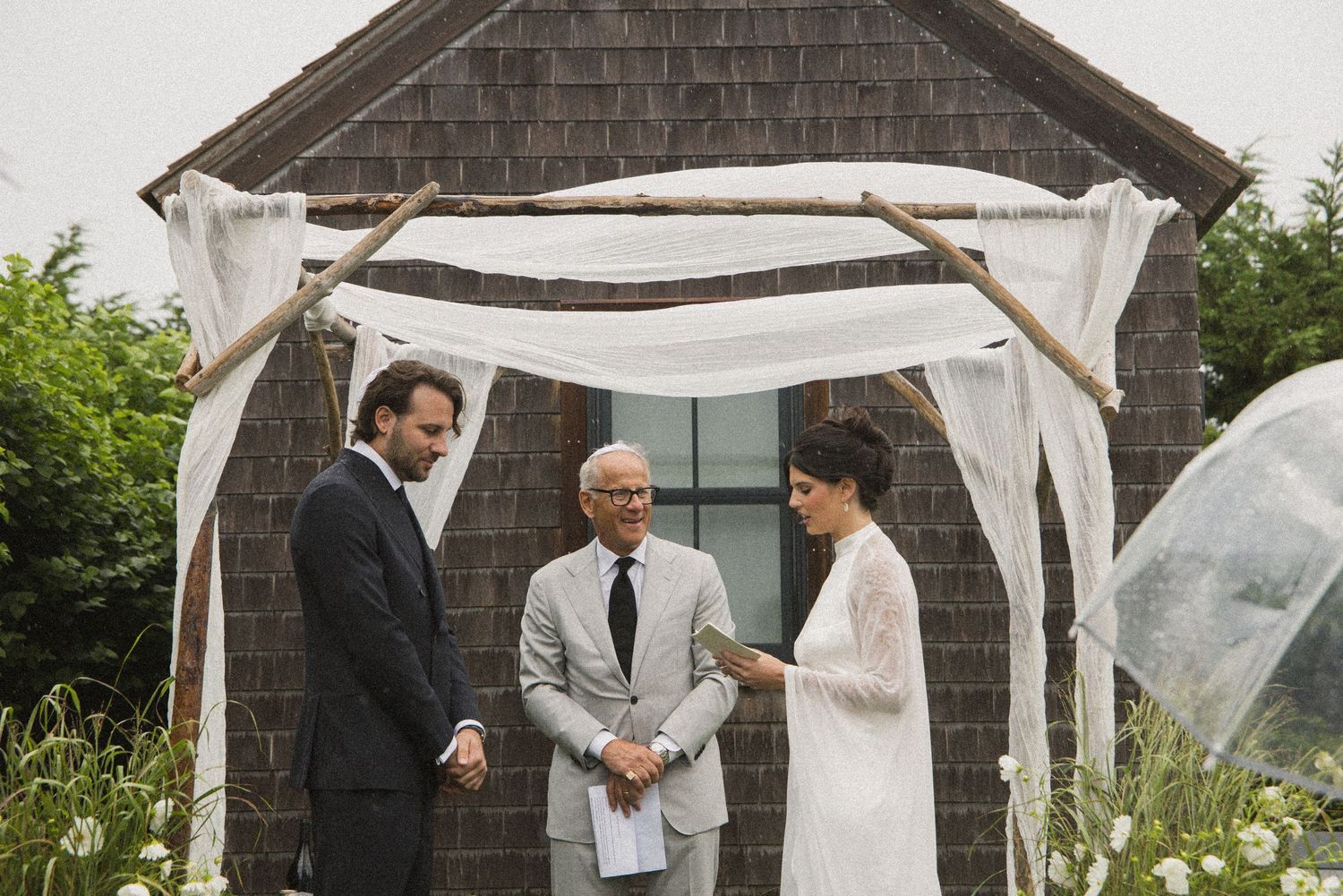 Couple exchanging vows outdoors under a rustic wooden chuppah draped with white fabric at a garden wedding.