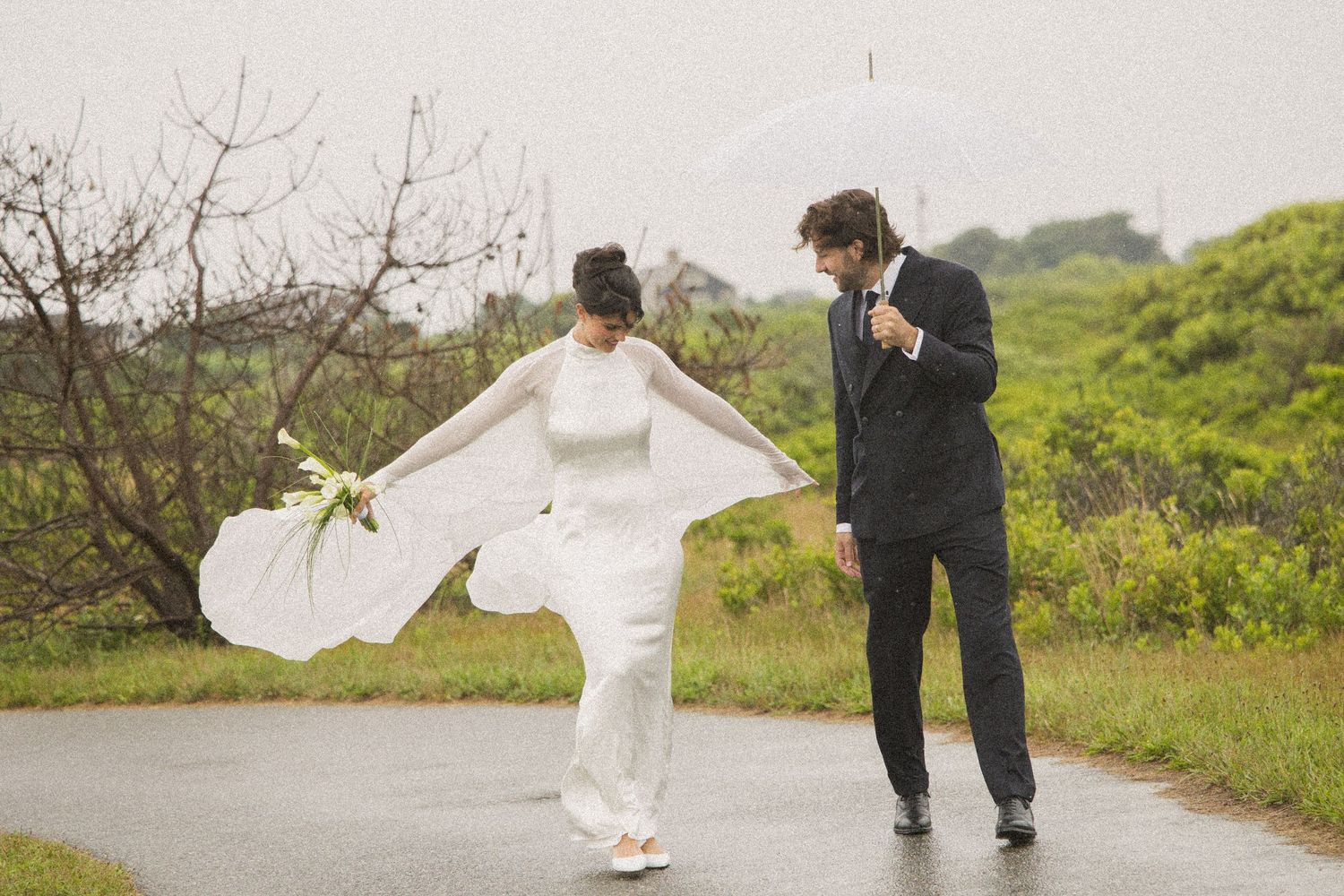 Bride and groom walking in rain under clear umbrella on wet road, bride's white gown flowing in wind.