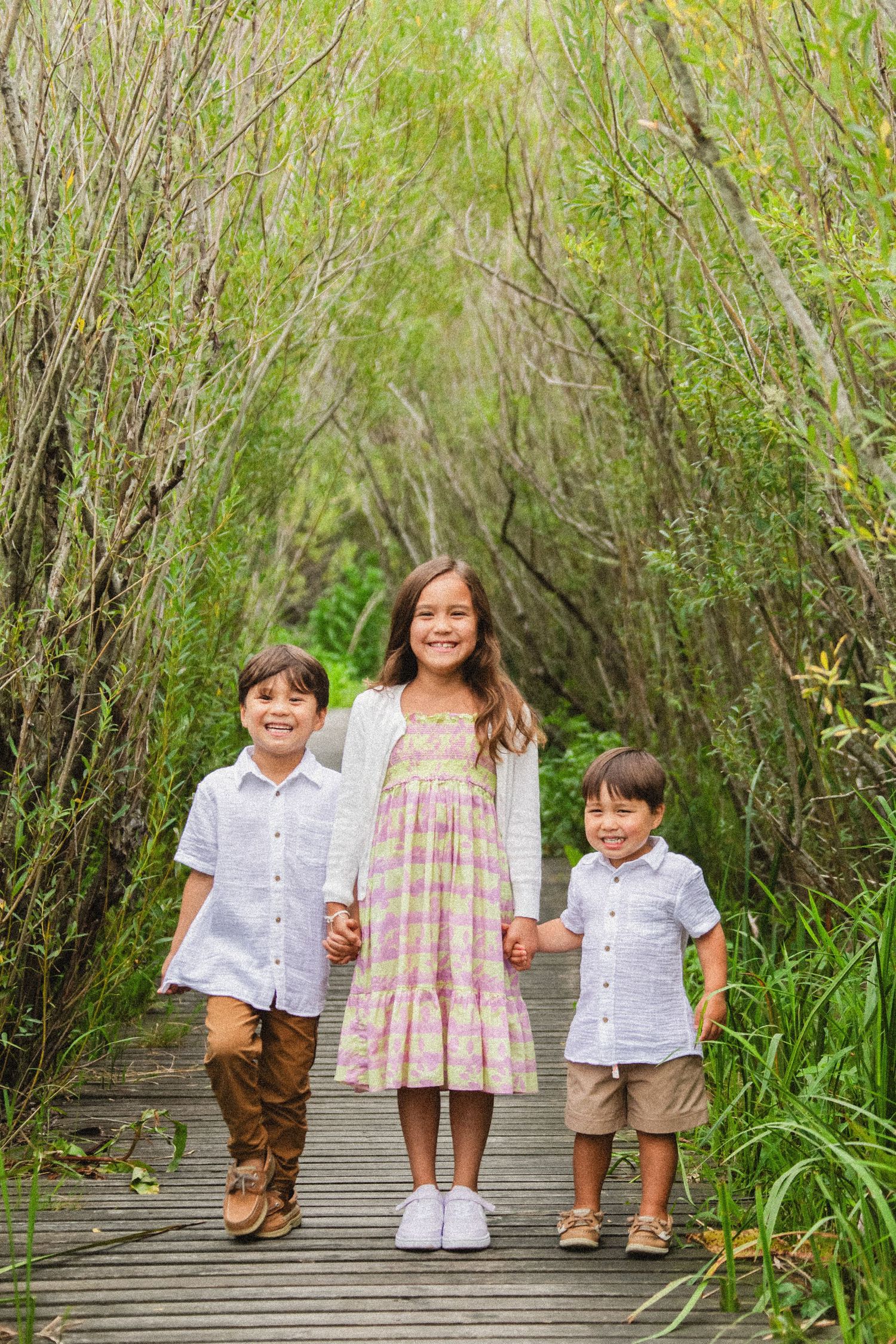 Three children hold hands while standing on a wooden boardwalk surrounded by green foliage.