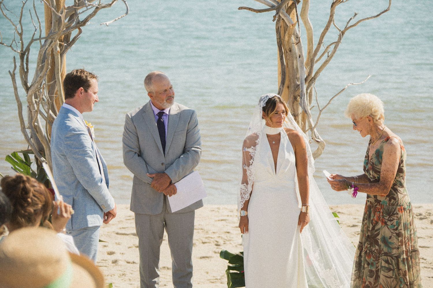 Wedding ceremony proceedings under driftwood arch on sunny beach.