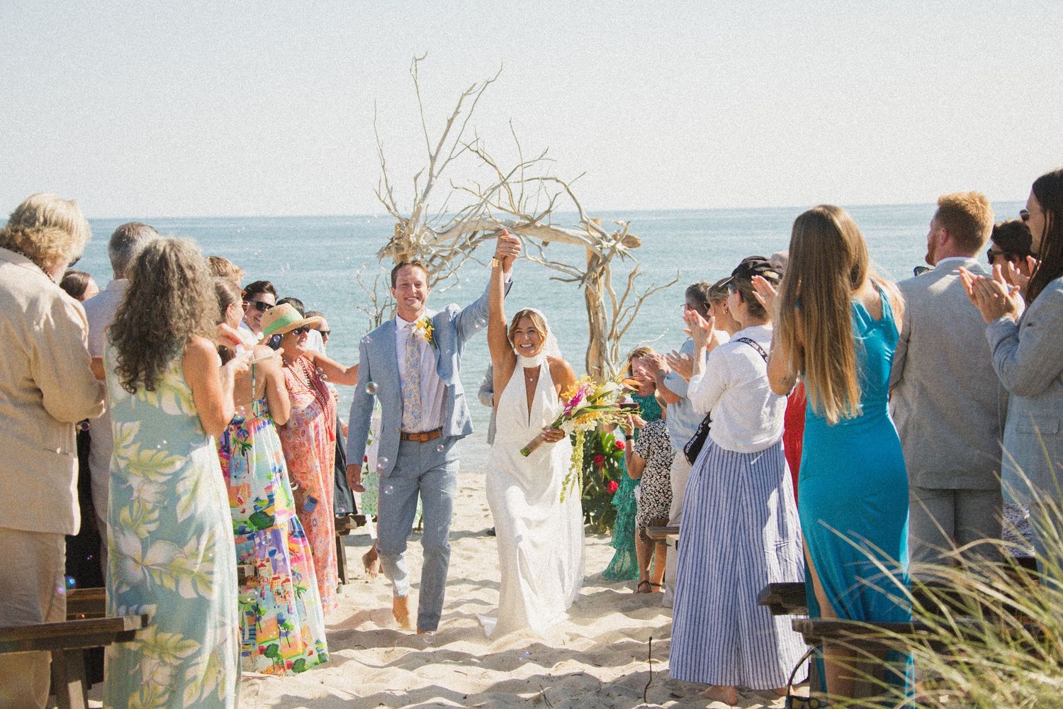 Wedding ceremony on a sunny beach with guests gathered around a driftwood arch overlooking the ocean.