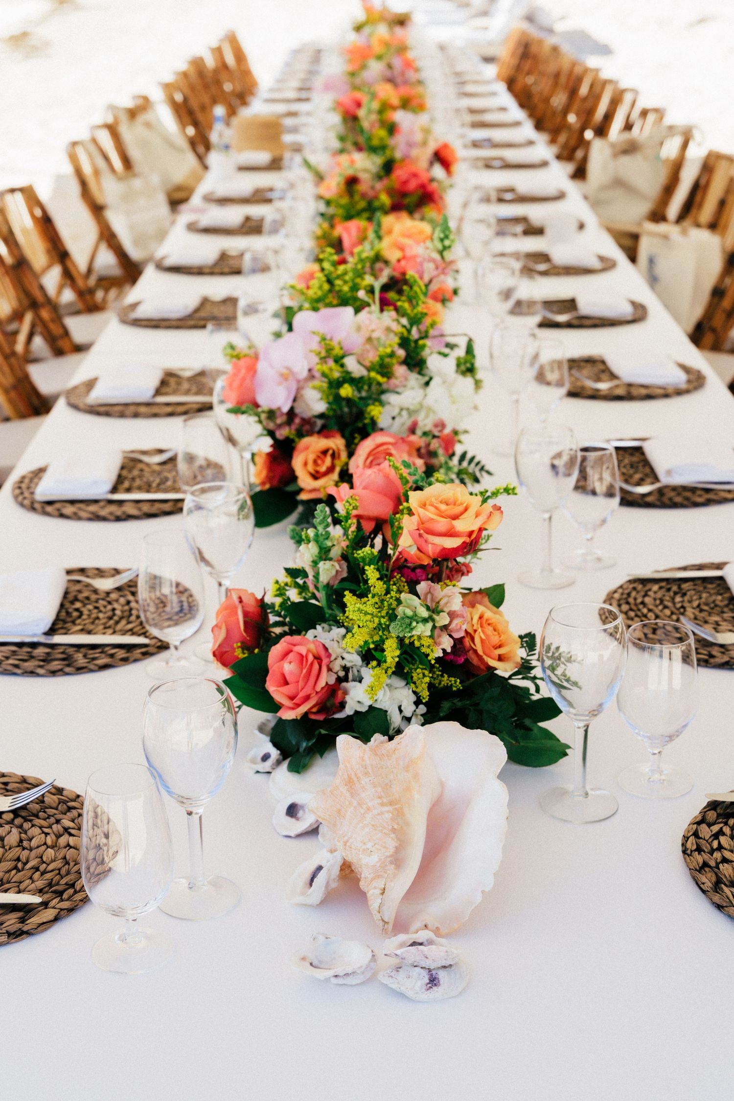 Long wedding reception table set with coral floral centerpieces, white linens, and woven charger plates.