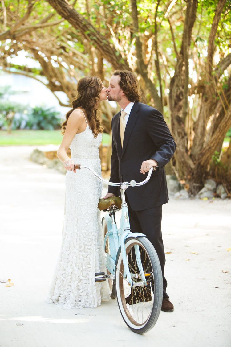 A couple in wedding attire share a romantic moment with a vintage bicycle on a beach path.