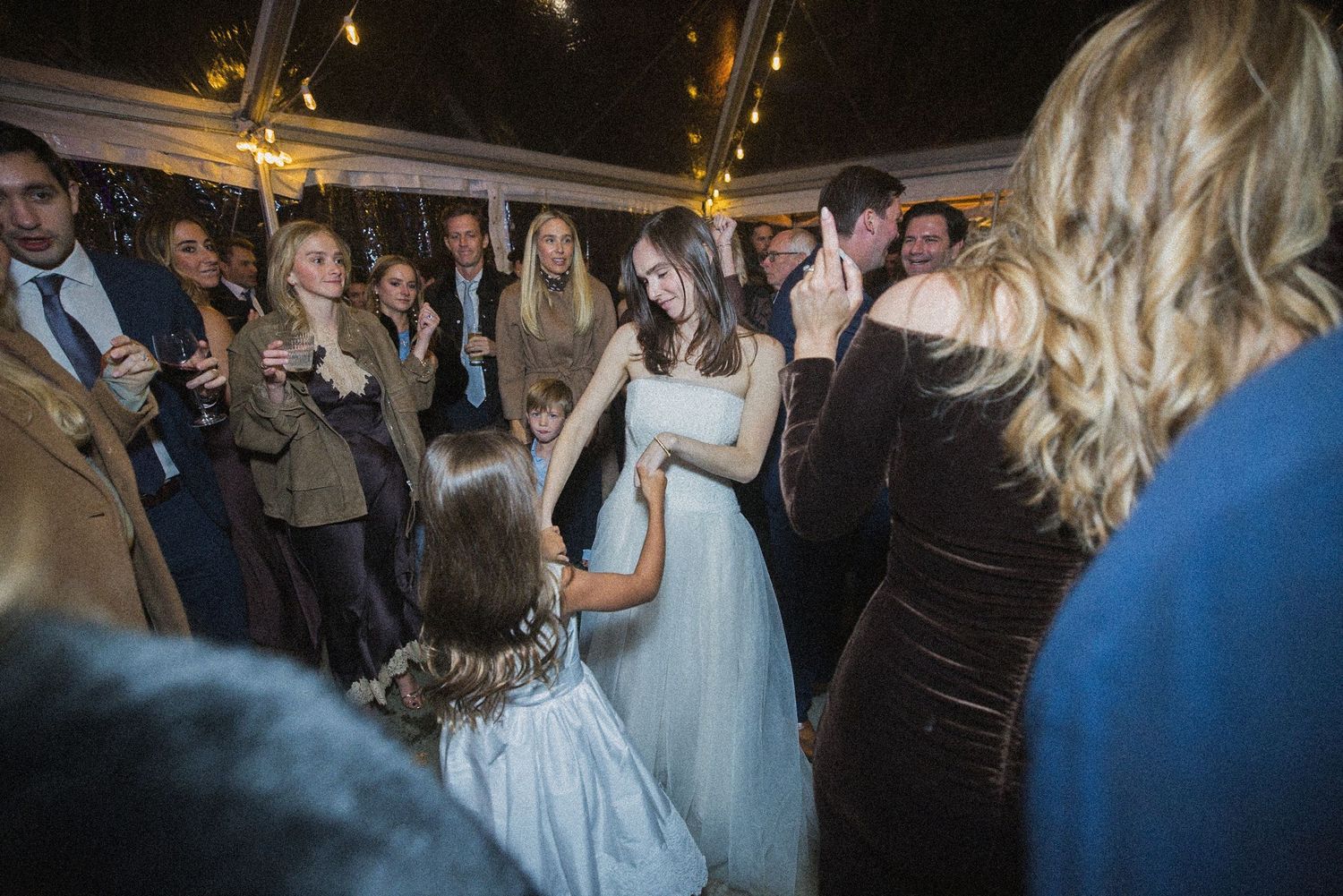 A bride in a white gown holds hands with a little girl in a flower girl dress as wedding guests gather around under a lit tent.