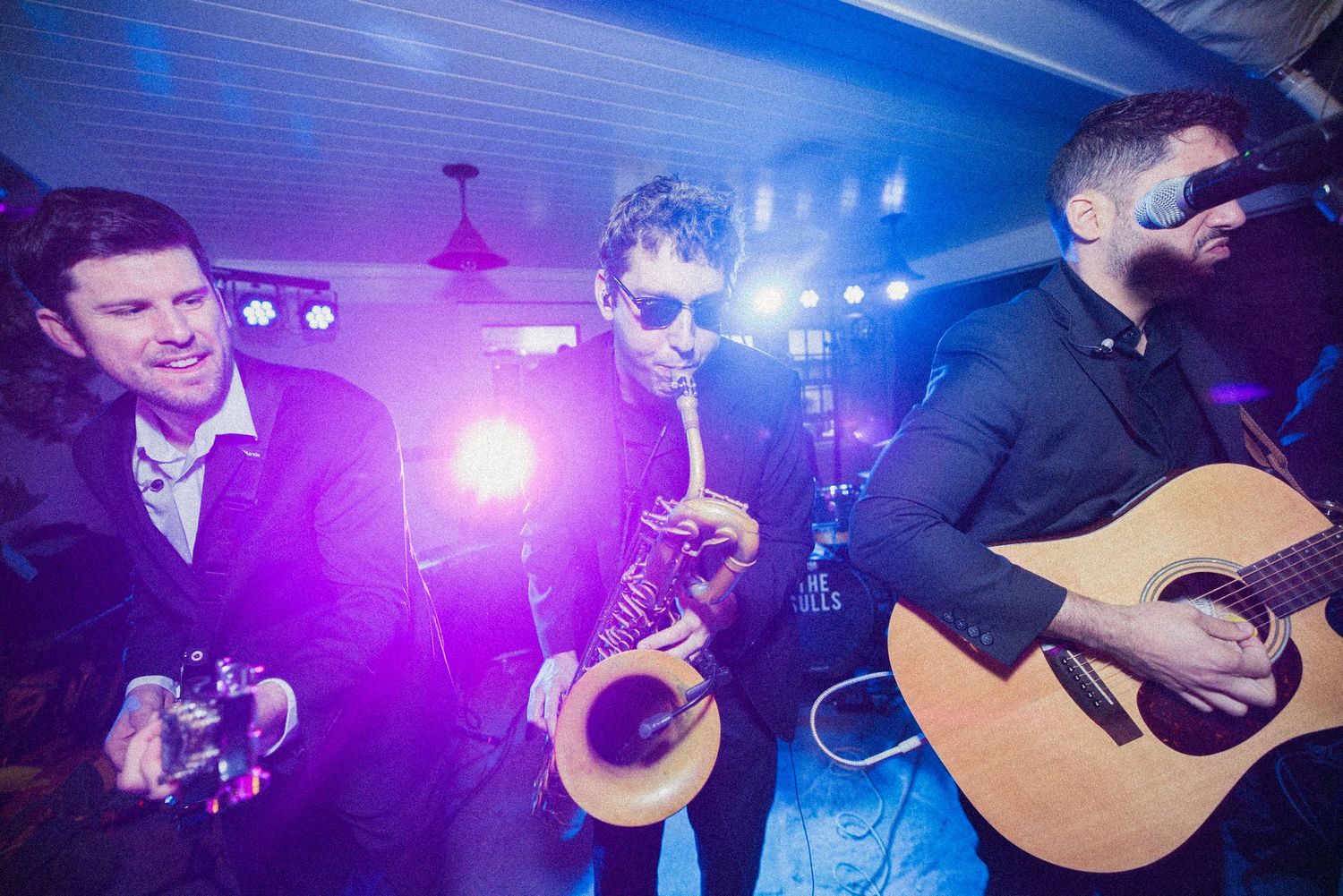 Three band members perform on stage under purple lights, with a guitarist, a saxophonist in sunglasses and a smiling musician with a camera.