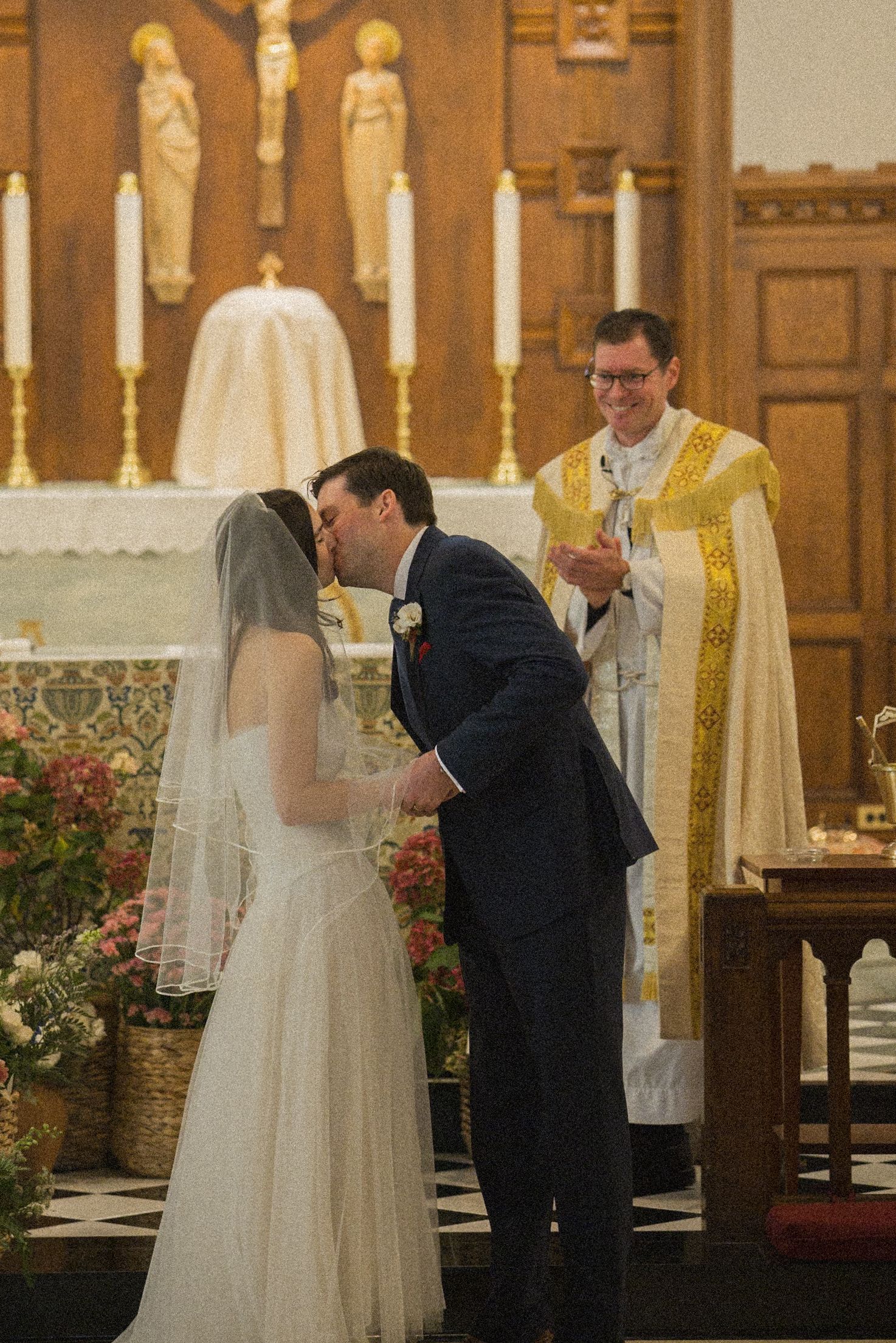 A bride and groom share their first kiss at the church altar as a smiling priest in gold vestments looks on.