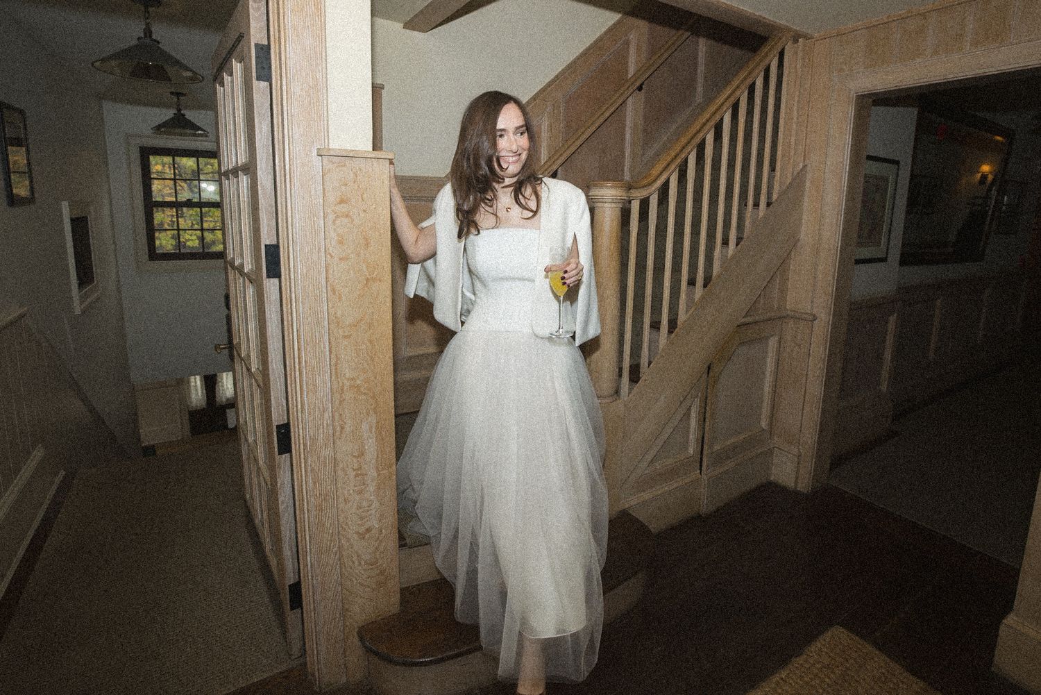 Smiling woman in a white tulle gown and shawl stands beside a wooden staircase in a rustic home, holding a drink.