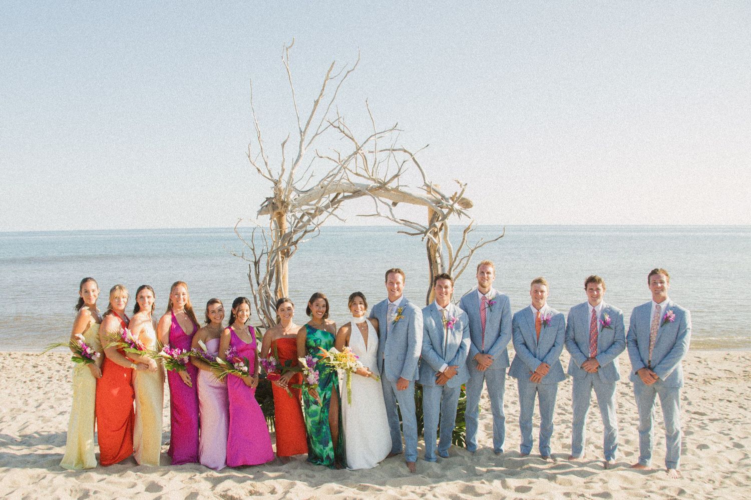Wedding party poses under driftwood arch on beach with bridesmaids in colorful dresses.