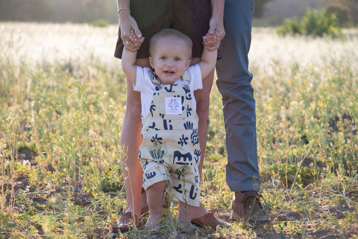 A smiling toddler learns to walk in a wildflower field at sunset.