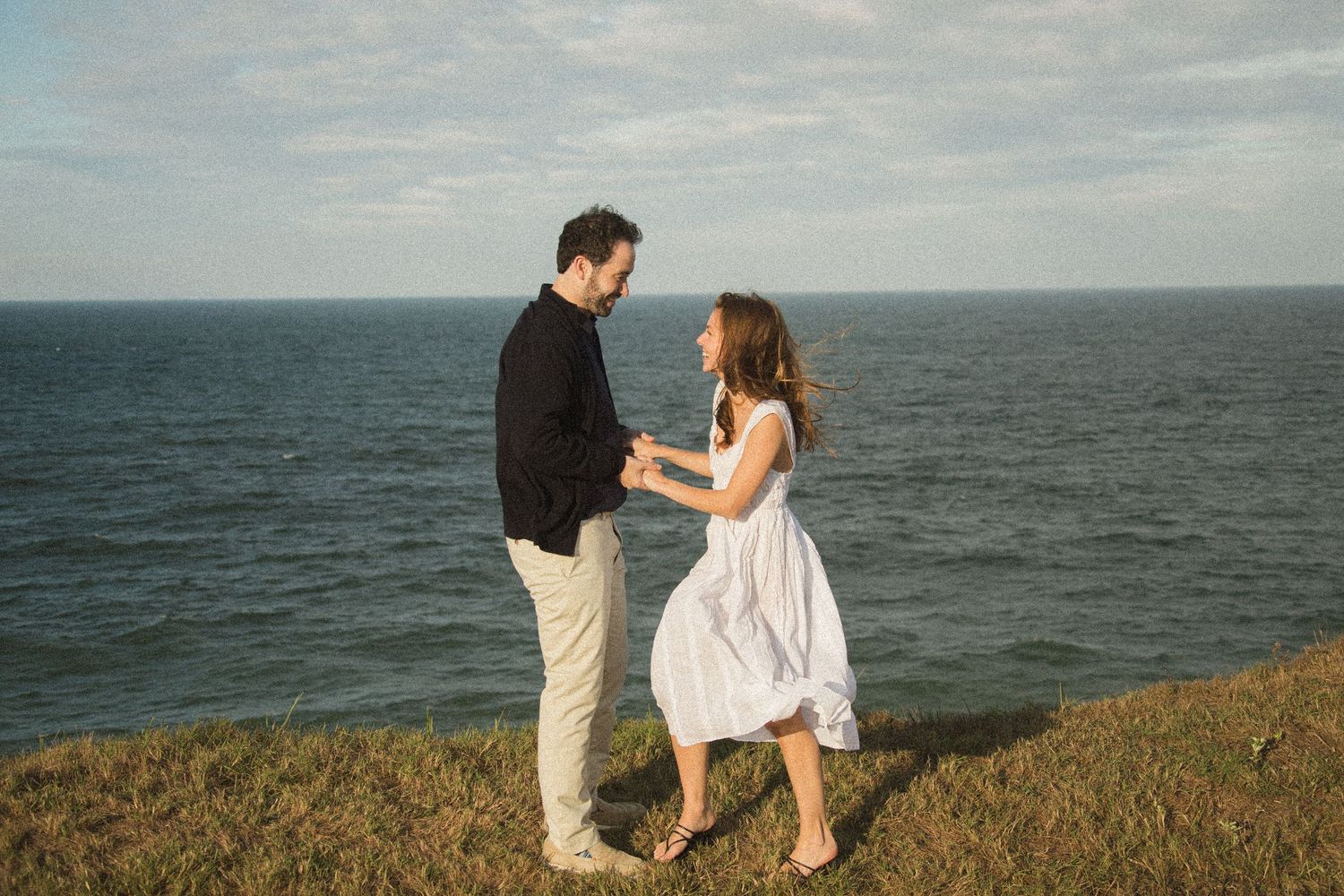 A couple dances on a coastal cliff overlooking the ocean at sunset.