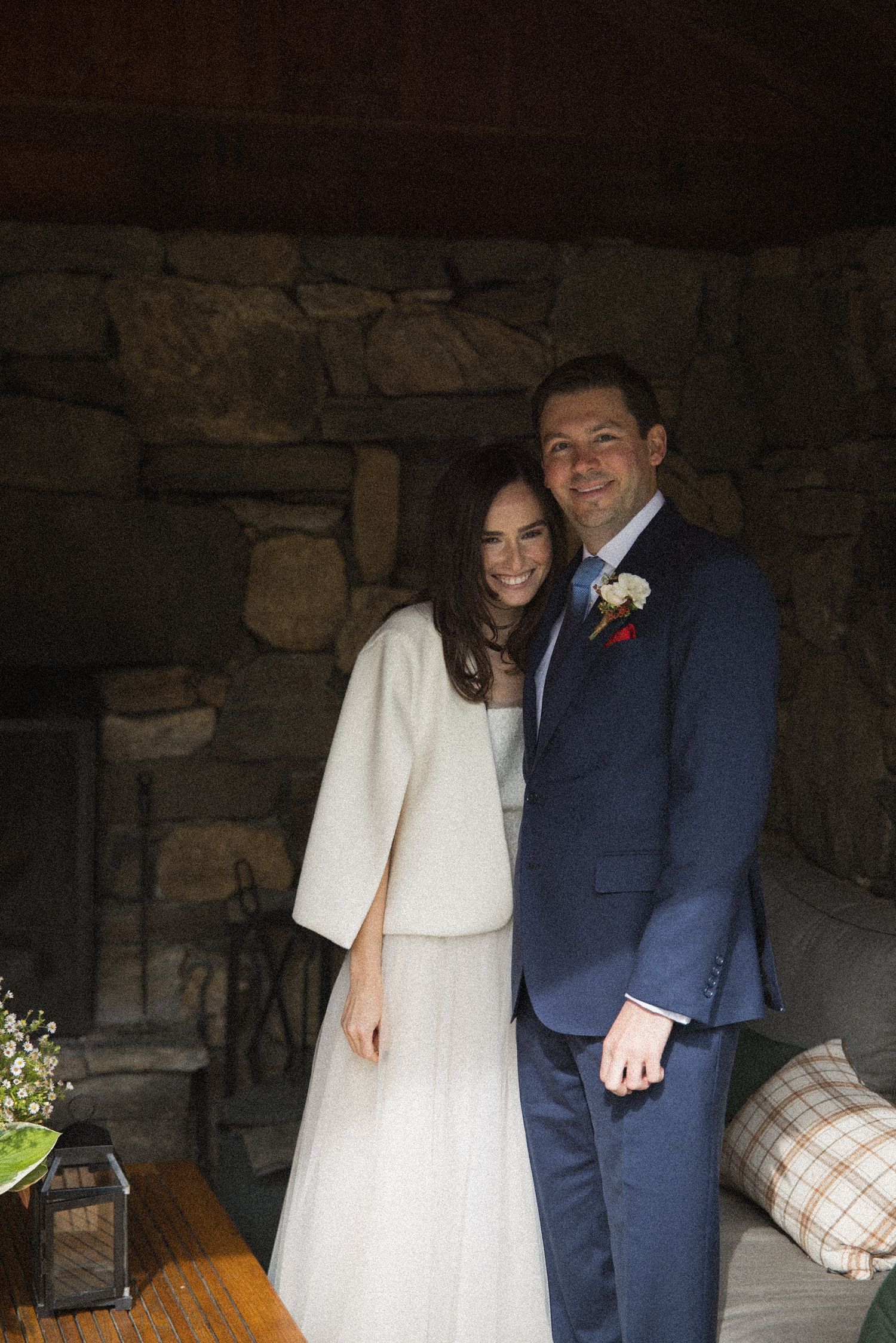 Smiling bride in a white skirt and ivory jacket stands beside a groom in a navy suit with a boutonniere before a rustic stone wall.