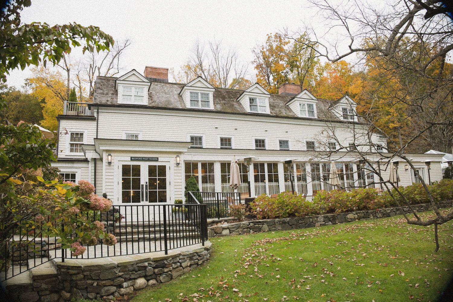 White colonial-style house with dormer windows, stone retaining wall, and a lush green lawn surrounded by colorful autumn trees.