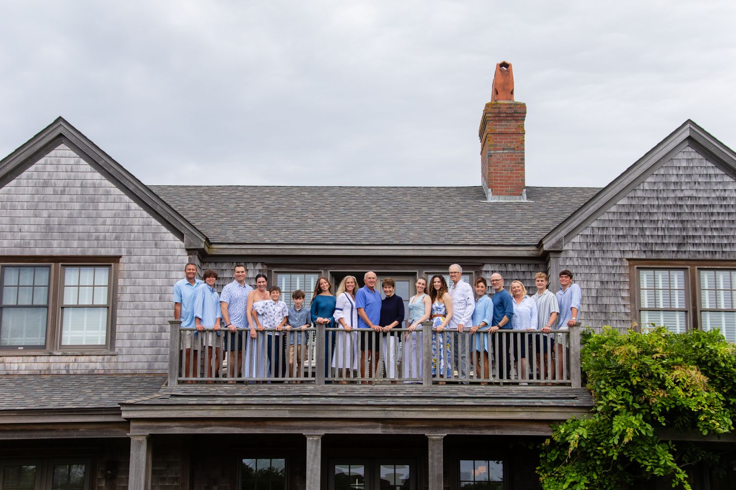 Group of people posing on a wooden deck balcony of a shingle-style coastal home under cloudy skies.