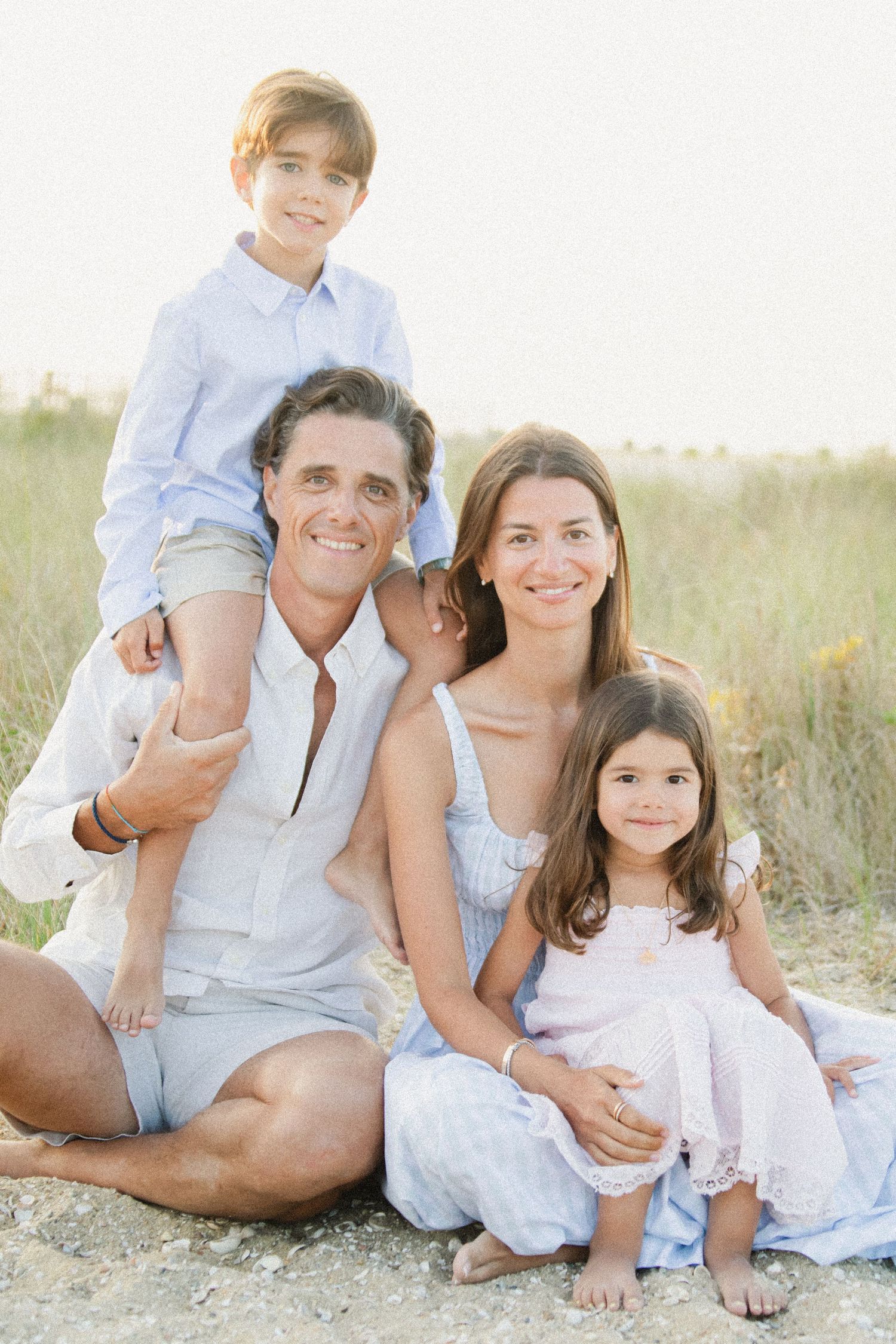 Happy family of four posing together on a sunny beach, parents with two young children smiling outdoors.