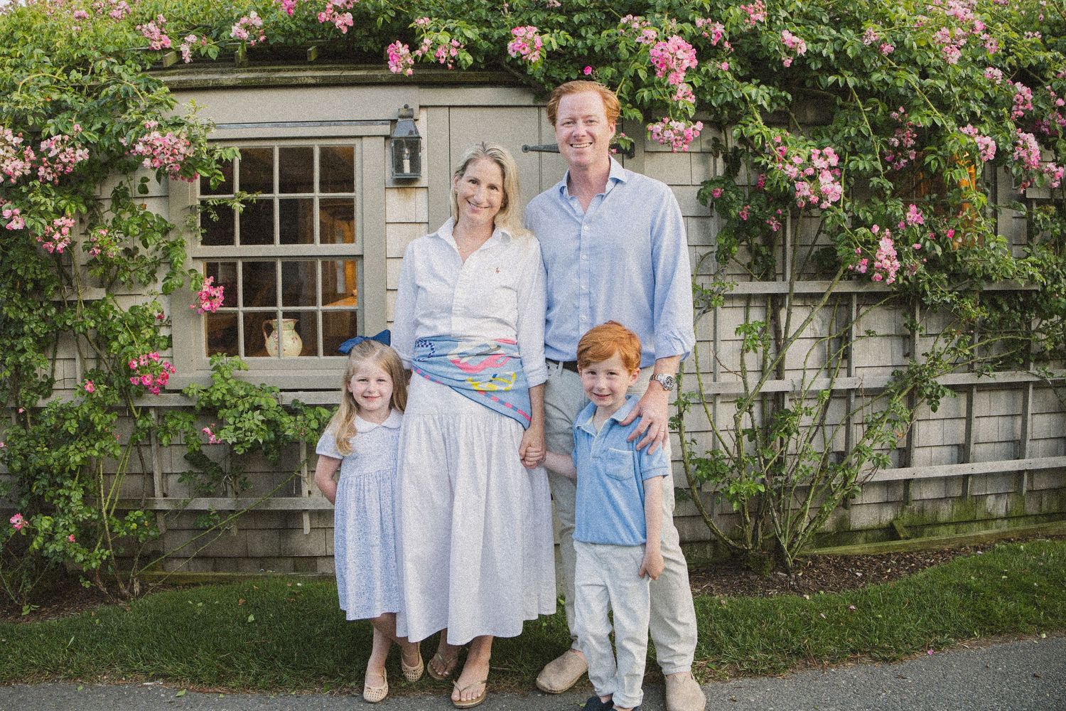 Family of four smiling in front of a charming cottage with blooming pink roses, dressed in coordinating blue and white outfits.