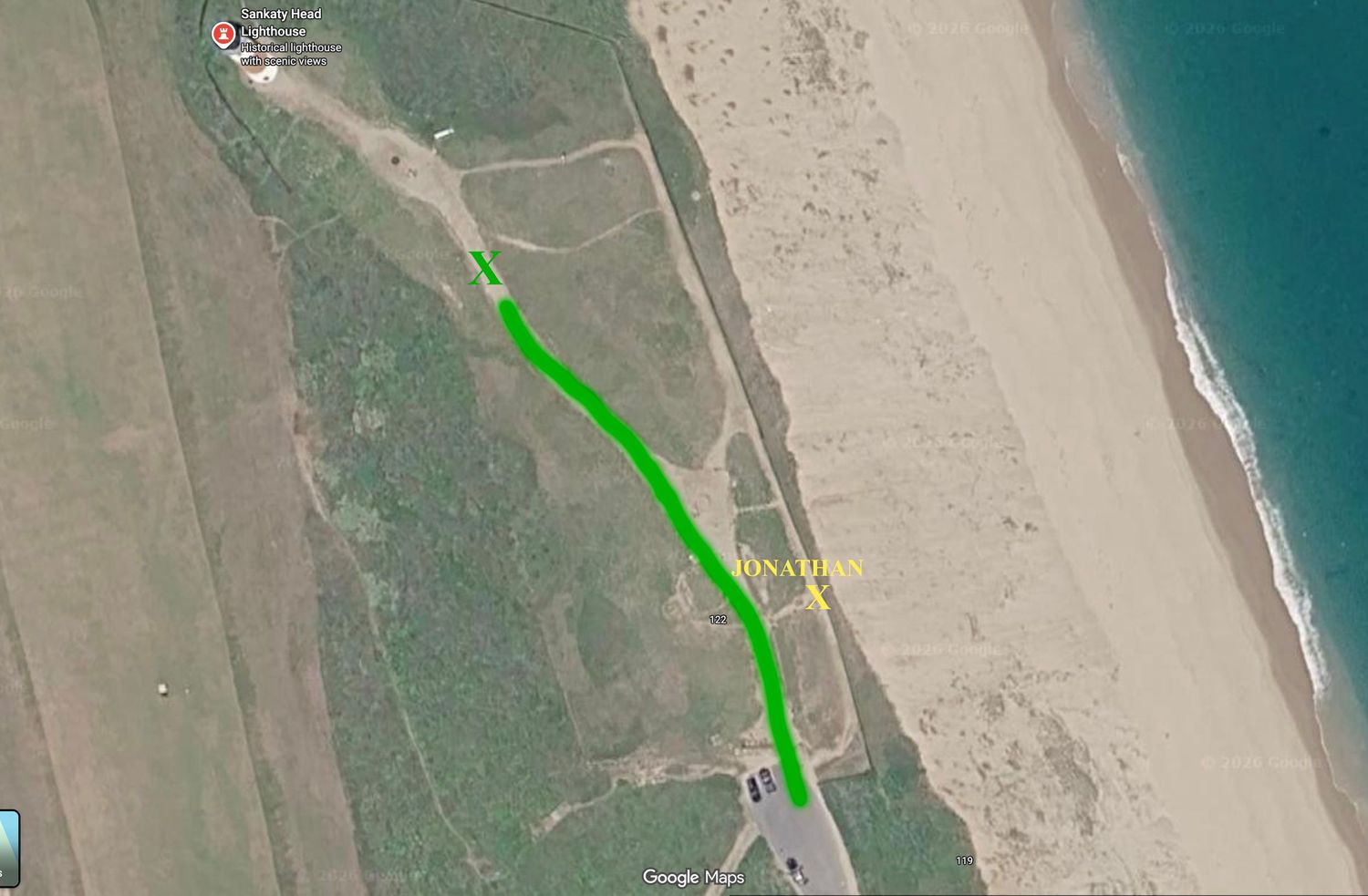 Aerial satellite view of a coastal path marked with a green route line along a sandy beach shoreline.