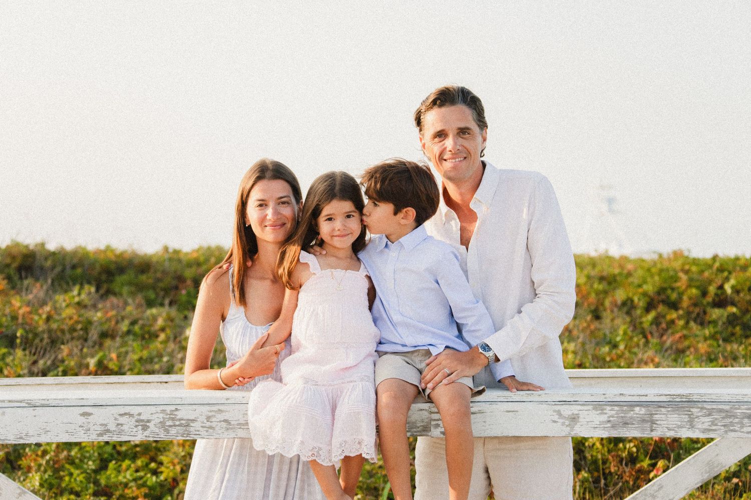 A happy family of four poses on a white fence at a sunny beach, wearing coordinated white and light blue outfits.