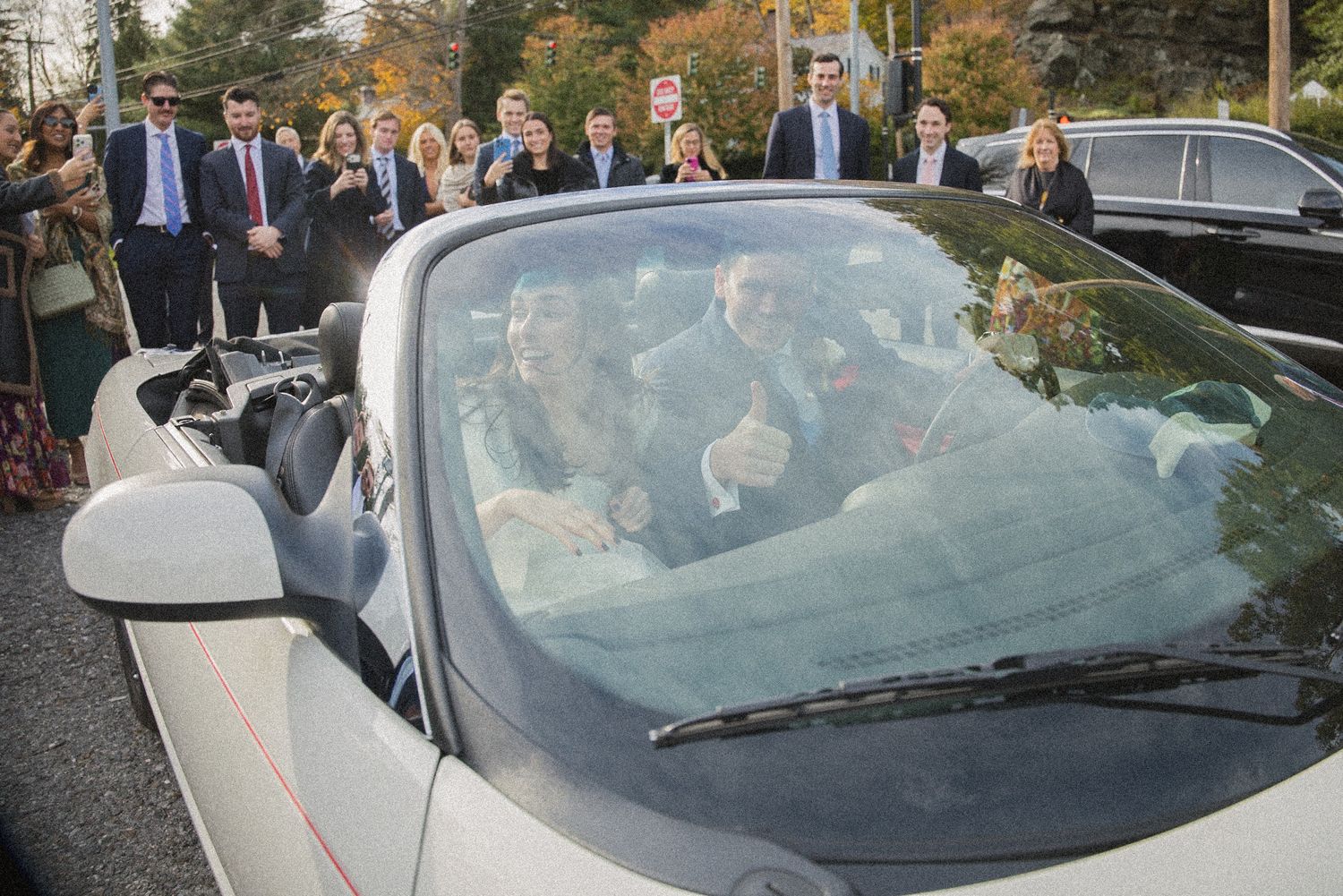 A newlywed couple waves from a white convertible as smiling wedding guests gather on the street behind them.
