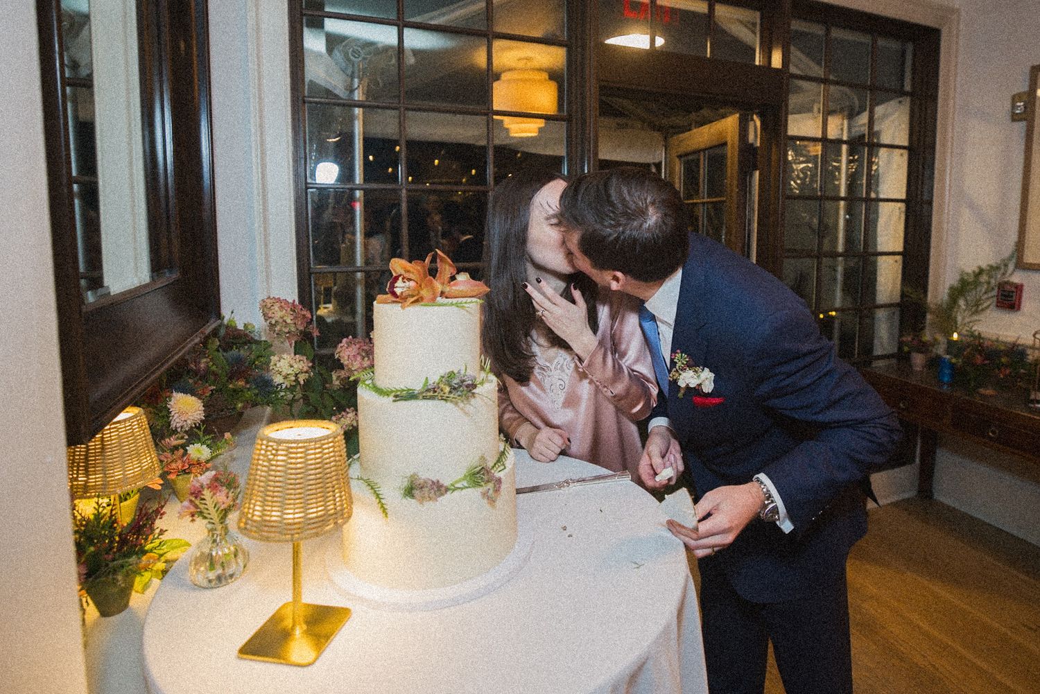 A bride and groom share a kiss beside a white tiered wedding cake decorated with fresh flowers and greenery.