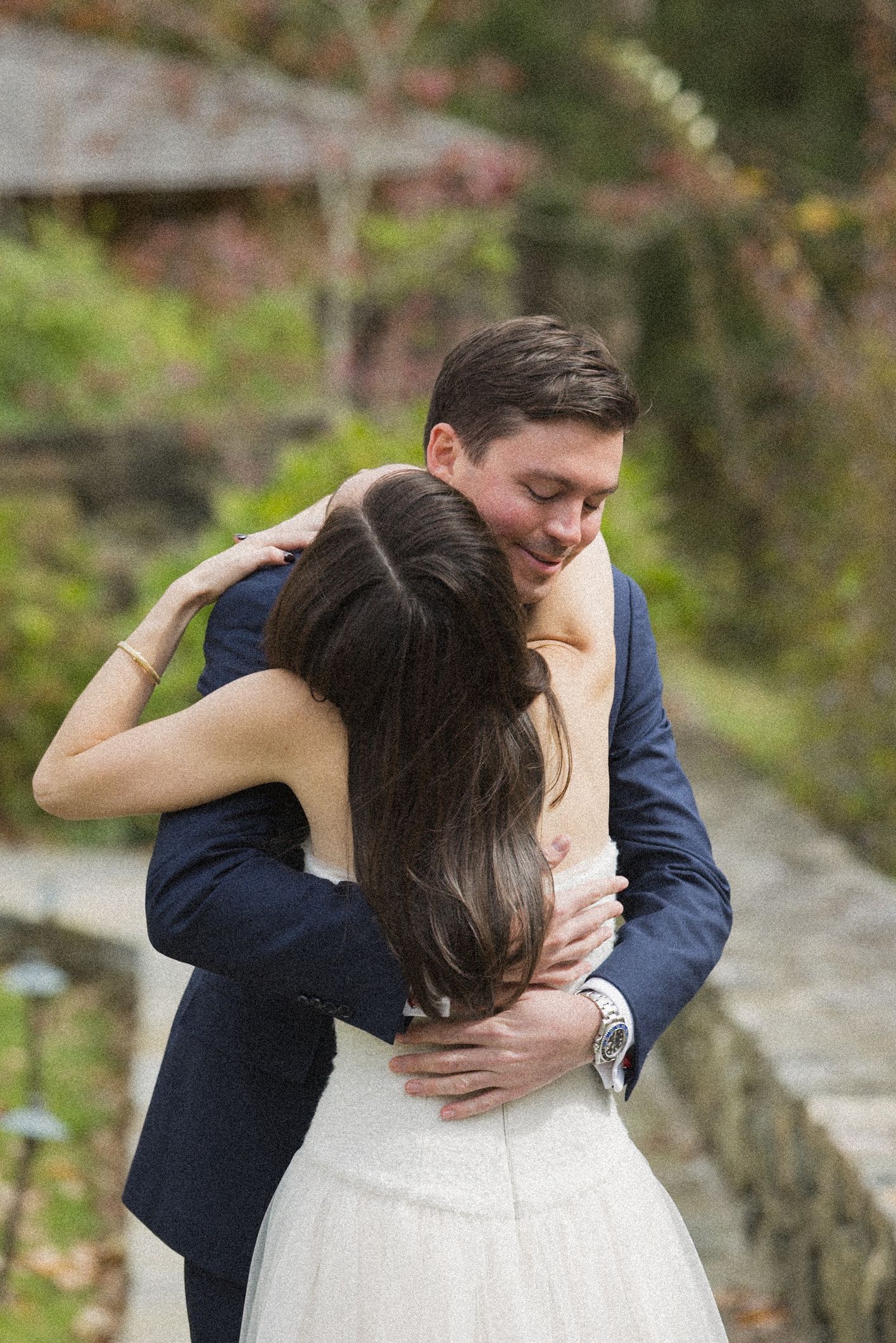 Groom in a navy suit embraces a bride in a white gown with long dark hair in a garden with autumn foliage.