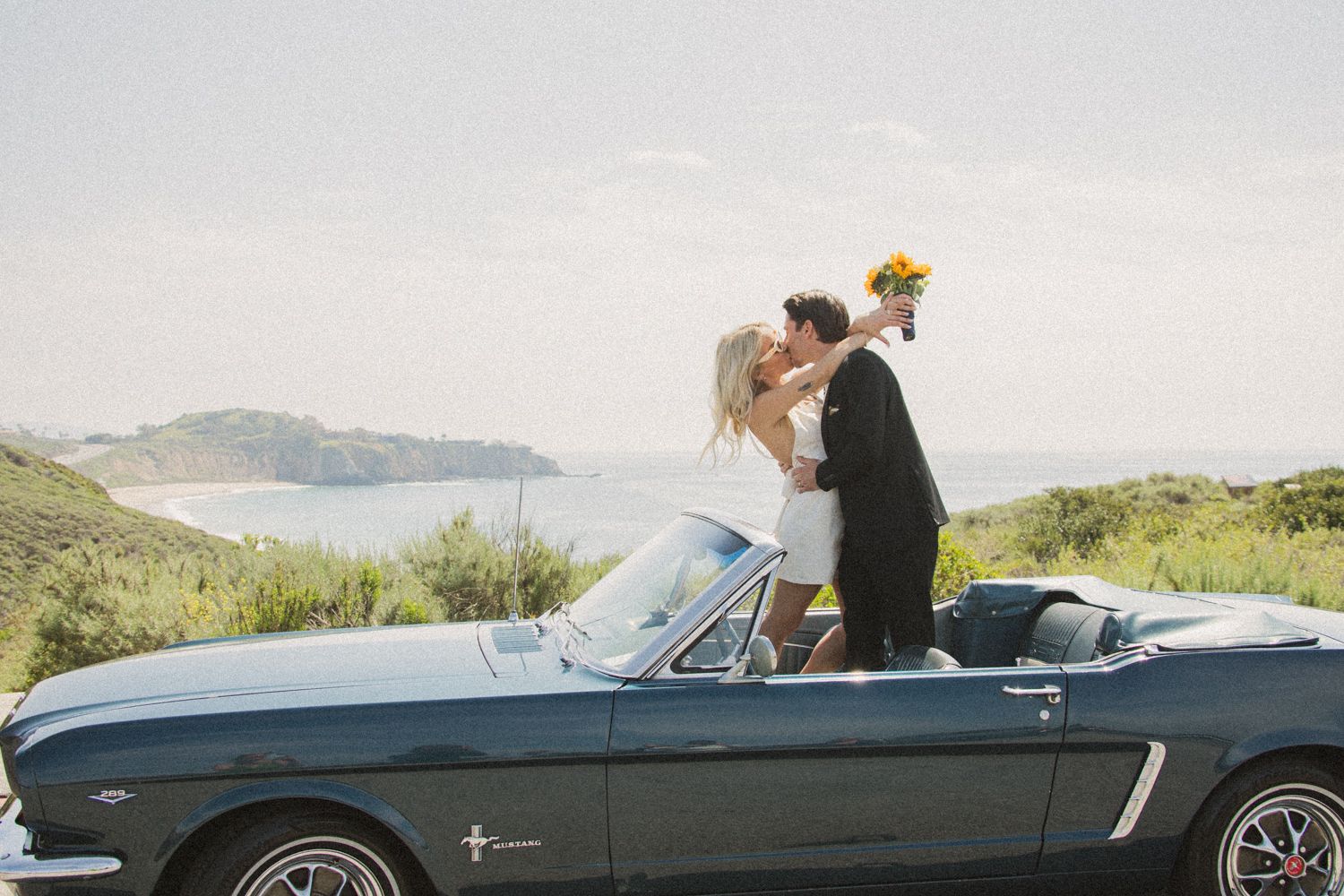 Classic convertible car driving along a scenic coastal road with mountains in the background.
