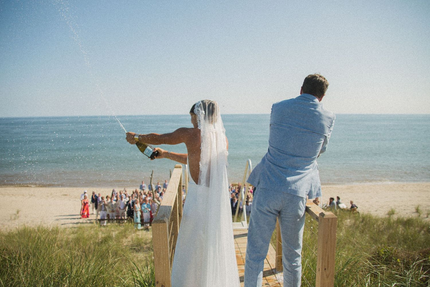 Couple stands on coastal boardwalk with wedding guests gathered on beach below.