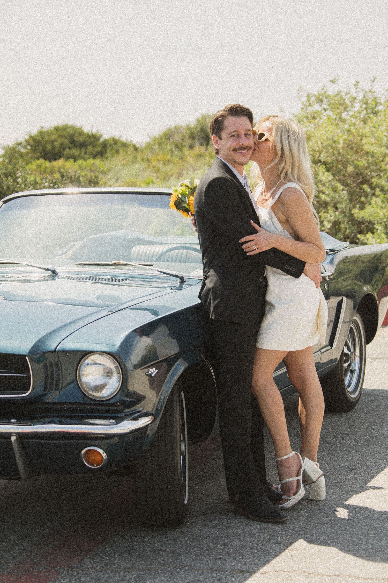 Romantic couple embracing against classic Mustang with mountains in background.