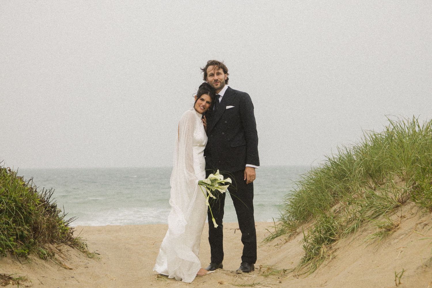 Couple in wedding attire posing on sandy beach dunes with ocean backdrop on a cloudy day.