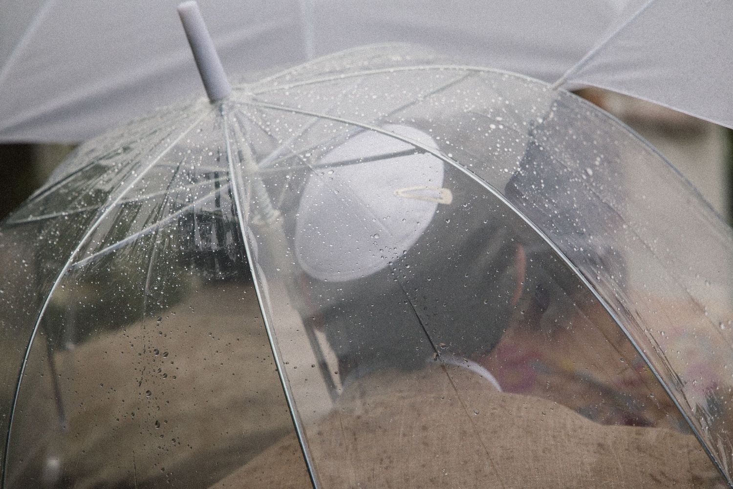 Person sheltering under a clear transparent bubble umbrella in the rain, with water droplets visible on the surface.