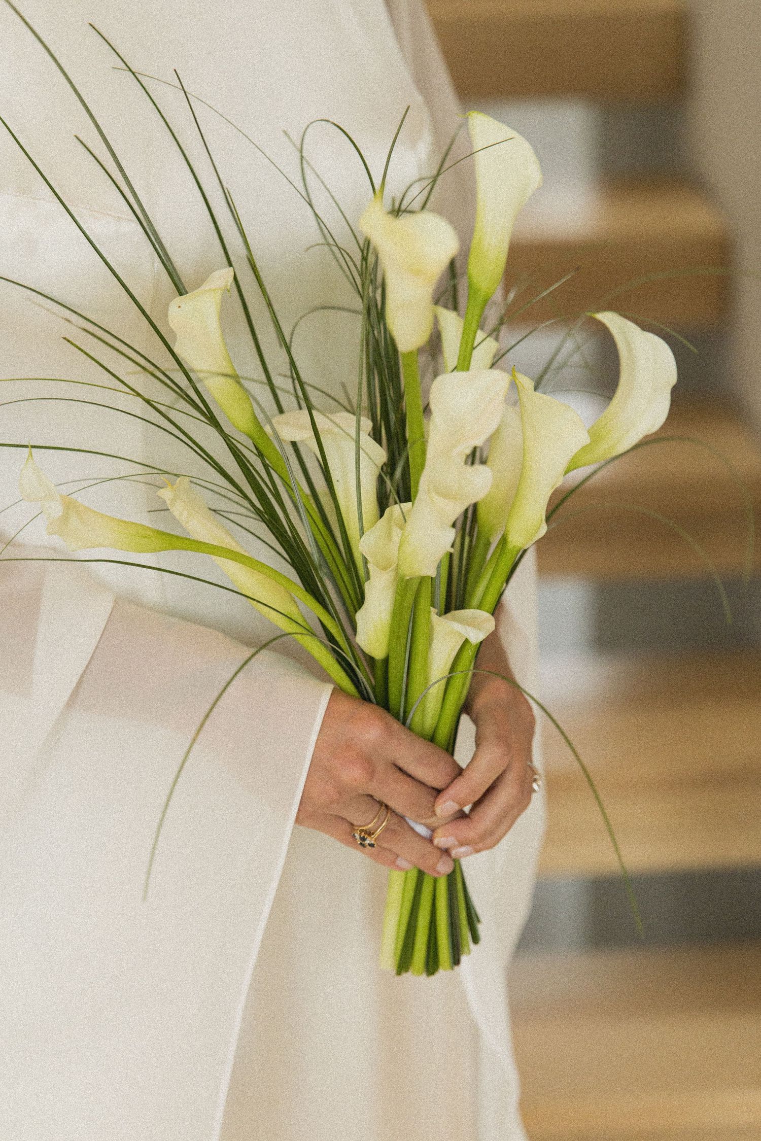 Bride holding elegant white calla lily wedding bouquet with green grass accents, wearing a white gown and gold ring.