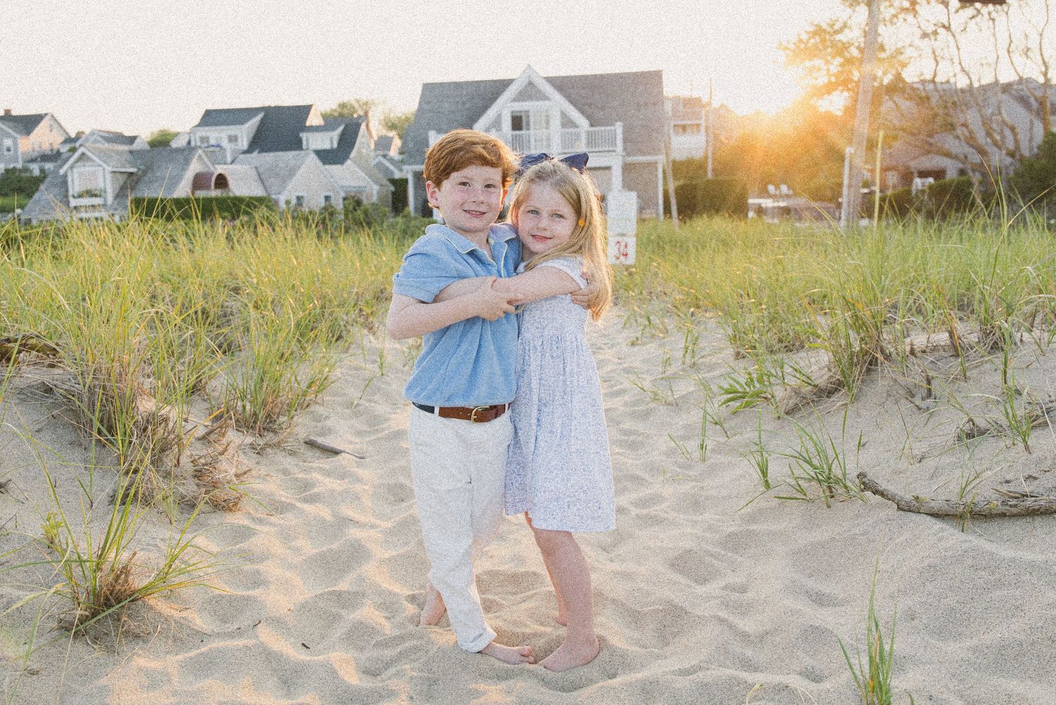 Two young children hugging on a sandy beach at sunset with coastal homes and sea grass in the background.