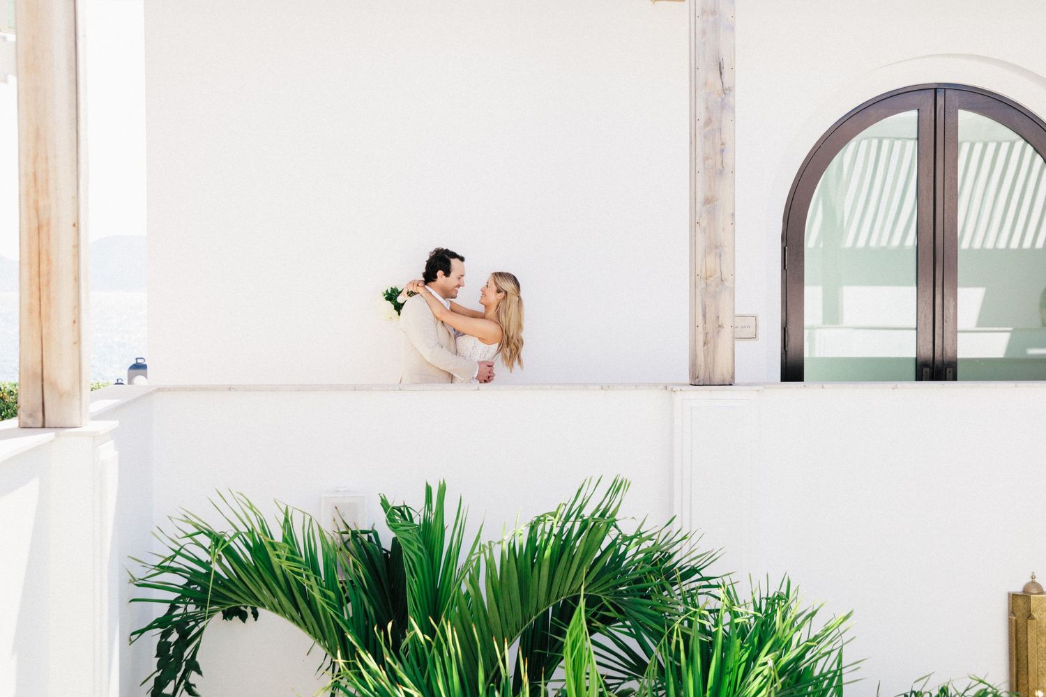 An intimate wedding moment captured on a white Mediterranean-style balcony with palm plants.
