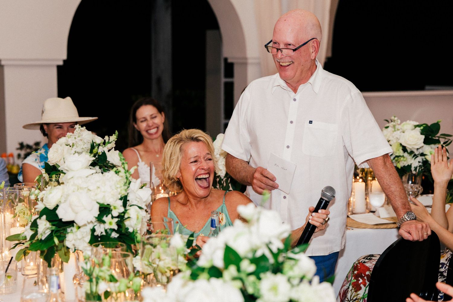 Wedding reception dinner with white floral centerpieces and guests seated at long tables.