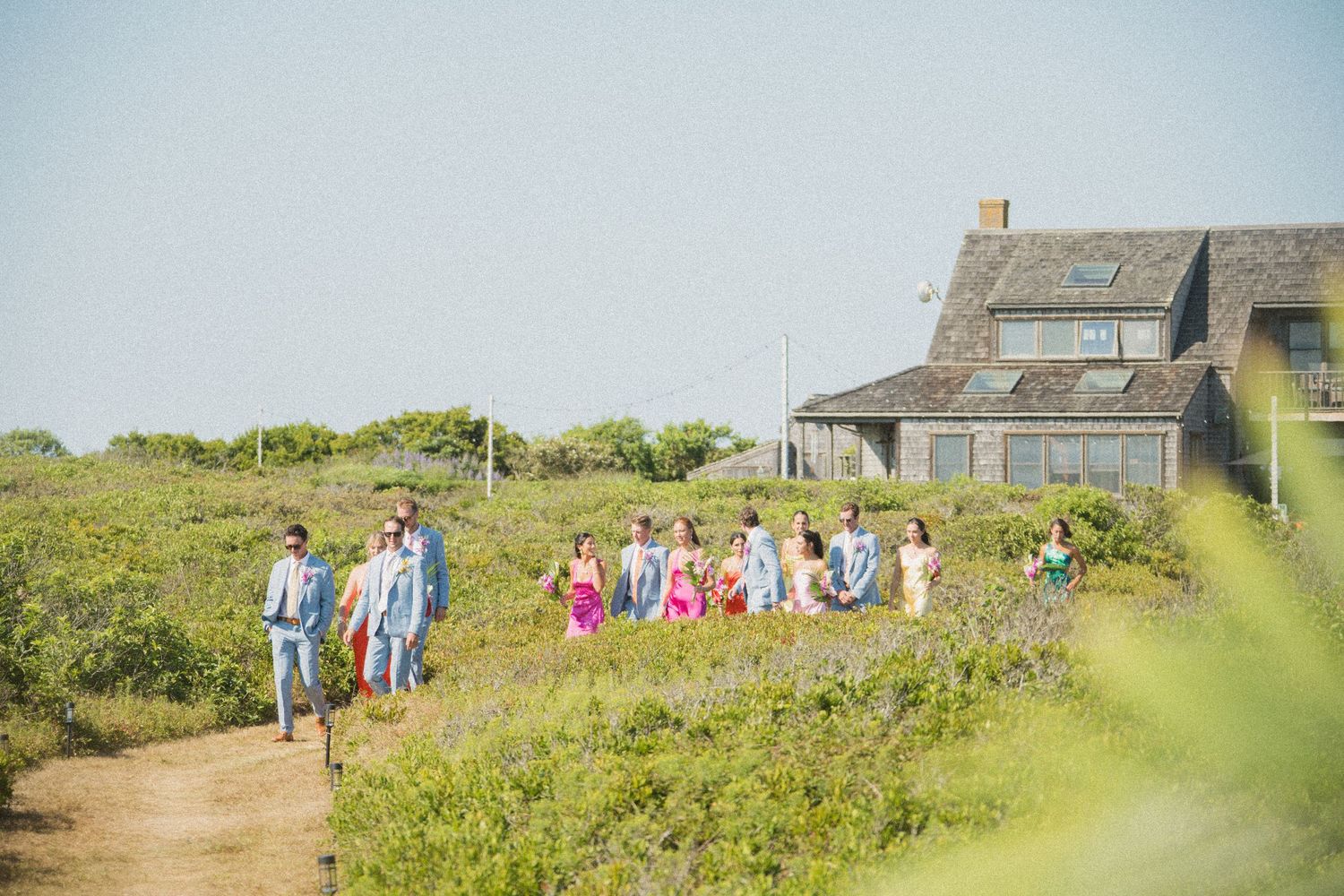Wedding party walking along coastal path with rustic buildings in background.