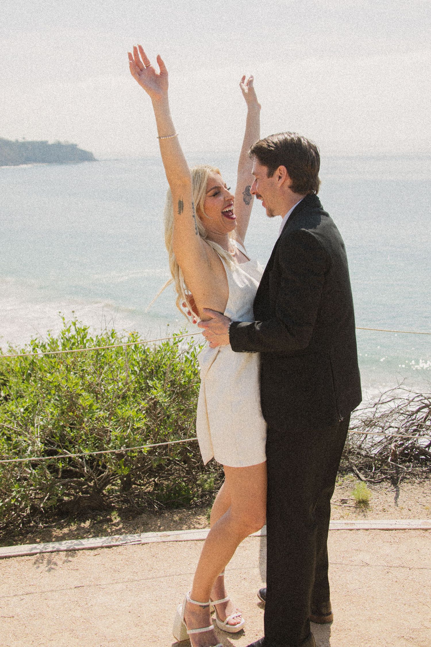 Newlyweds celebrate with raised arms on scenic coastal cliff after ceremony.