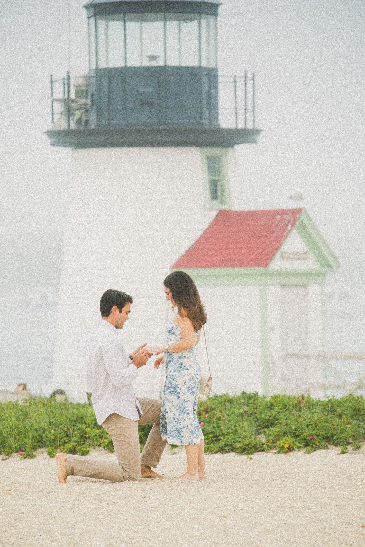 Man proposes to woman on sandy beach with historic white lighthouse and red-roofed building in background.
