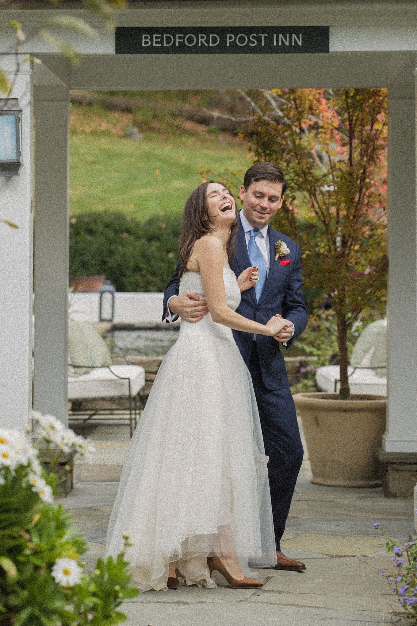 Laughing bride in a flowing white gown dances with her groom in a navy suit under the entrance of the Bedford Post Inn.
