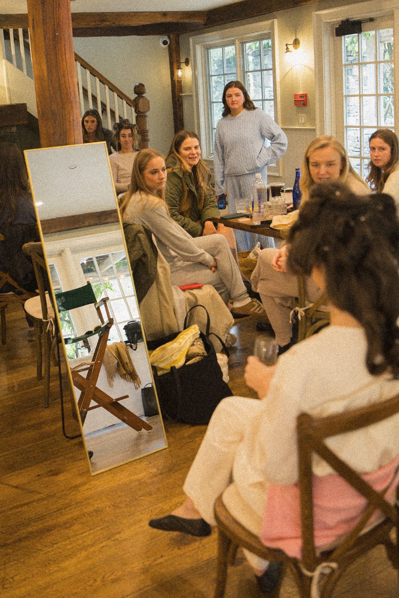 Group of women in casual outfits sit and stand in a sunlit room with wood floors, chatting beside a tall mirror before a celebration.