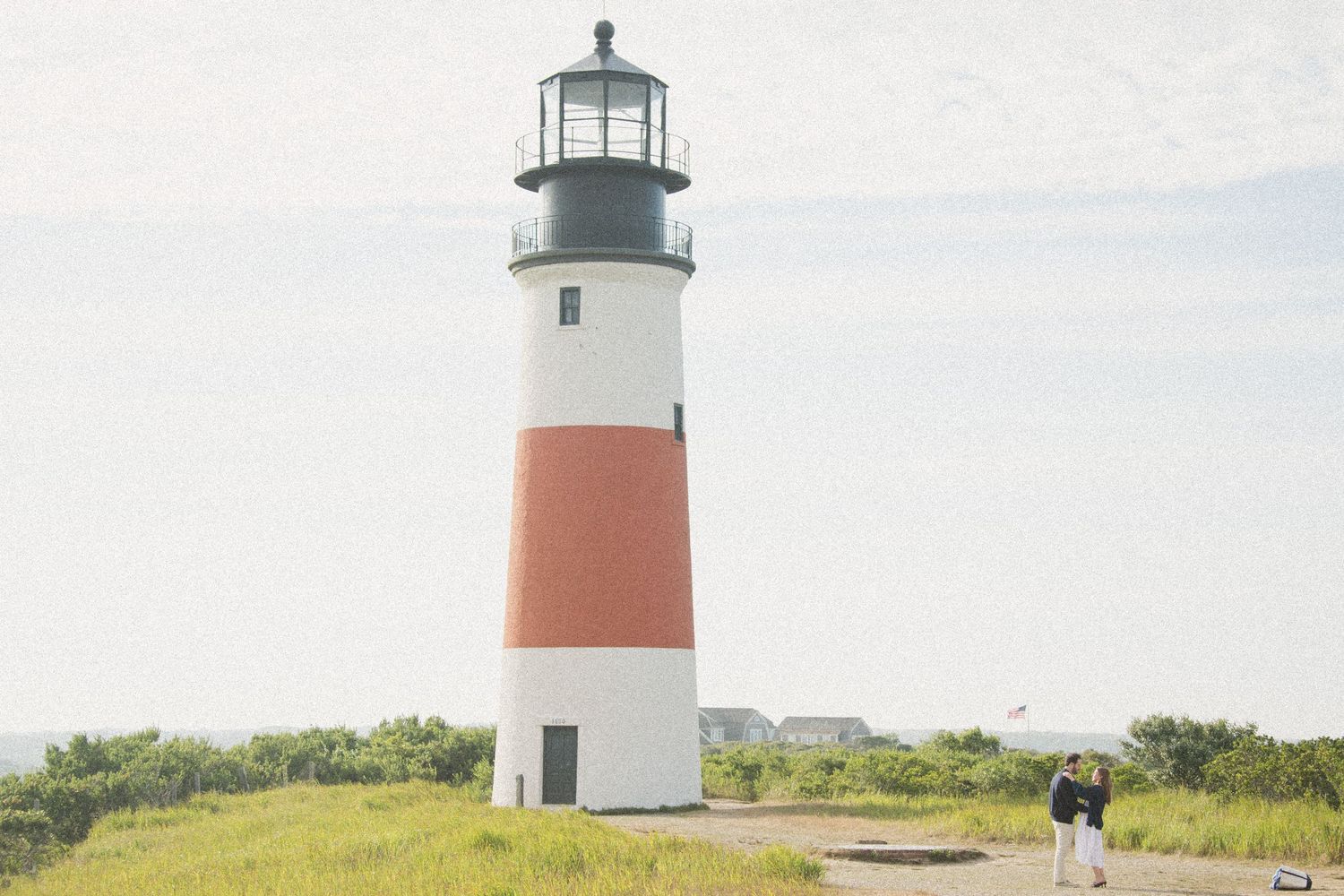 Lighthouse with red and white stripes stands against blue sky on grassy coastal landscape.