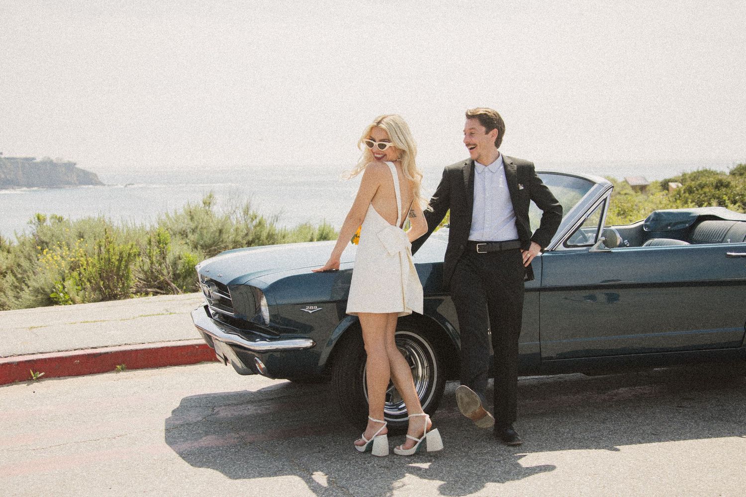 Couple sharing intimate moment next to vintage black Mustang convertible in scenic location.
