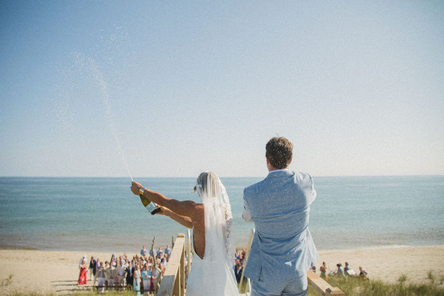 Newlyweds celebrate on elevated boardwalk overlooking beach crowd below.
