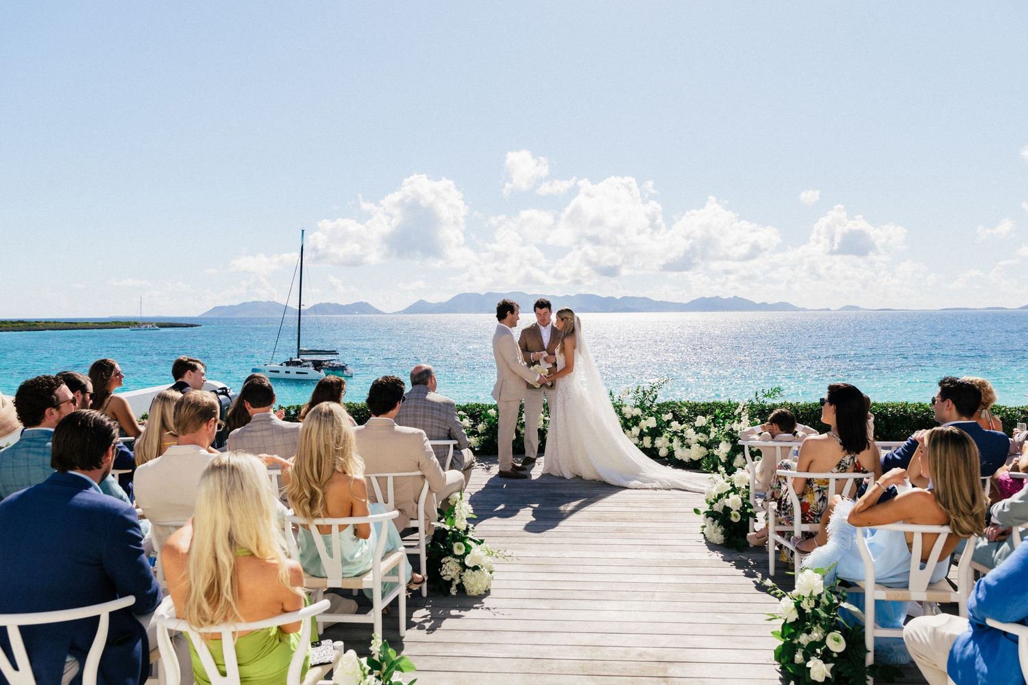 Panoramic view of a beach wedding ceremony setup with guests seated along a decorated wooden walkway.
