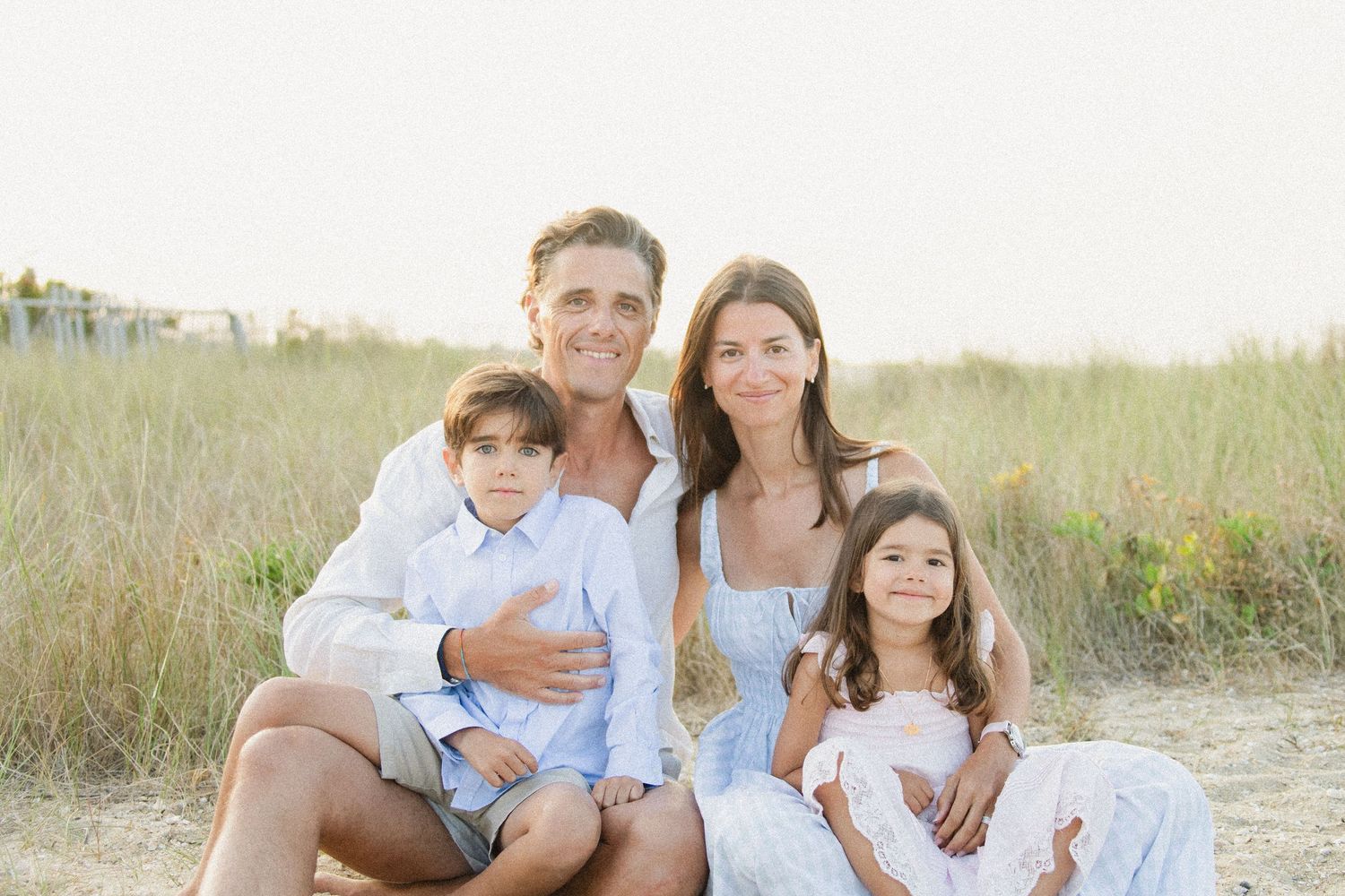 Happy family of four posing together on a sandy beach with green dunes in warm natural light.